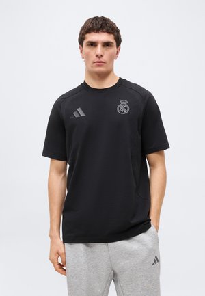 Muškarac u crnoj Real Madrid majici i svijetlosivim Adidas trenirking hlačama stoji ispred jednostavne pozadine.