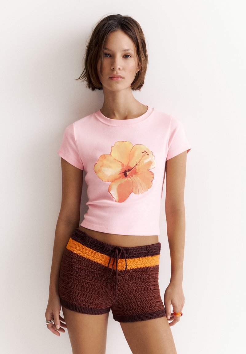 Jeune femme aux cheveux courts et bruns porte un crop-top rose avec une grande fleur orange et un short en maille marron avec des rayures orange.