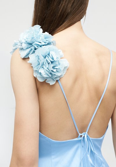 Γυναίκα με ανοιχτό μπλε σατέν φόρεμα με λεπτές τιράντες και δύο μεγάλα μπλε υφασμάτινα λουλούδια στον αριστερό της ώμο, με γυμνή πλάτη.
