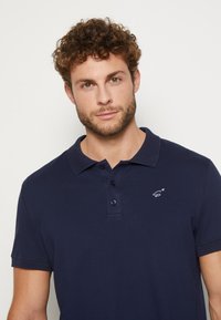 Navyblaues Poloshirt mit Kragen, drei Knöpfen und einem kleinen weißen Logo auf der linken Brust, aus einem glatten, strukturierten Stoff gefertigt.