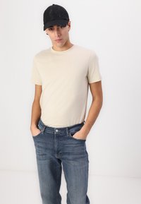 T-shirt en coton beige à manches courtes, associé à un jean en denim bleu. Modèle portant une casquette noire, les mains dans les poches, fond neutre.