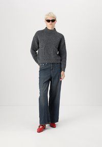 Grijze, gestructureerde gebreide trui met een hoge kraag en voorzakken, gecombineerd met losse blauwe spijkerbroeken en rode ballerina's.