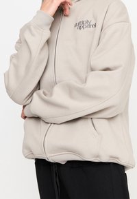 Sudadera de color beige claro con cremallera, bolsillos tipo canguro, puños acanalados y logotipo bordado en el pecho. Textura suave, corte relajado.
