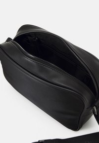 Černá kožená crossbody kabelka s horním zipovým uzávěrem, hladkou texturou a vnější kapsou. Disponuje odnímatelným popruhem pro univerzální nošení.