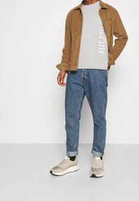 Brun kordfløjljakke, grå grafisk t-shirt, blå denim jeans og beige sneakers med creme detaljer. Opfoldede ærmer på jeans.