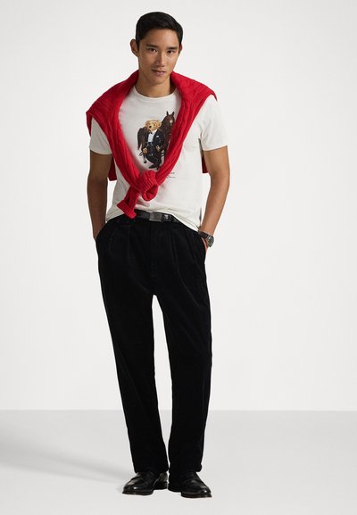 Valge T-särk graafilise printimisega, mustad puuvillased püksid ja punane kudumiseeniv kampsun, mis on hõlmatud õlgade ümber. Mustad jalanõud ja kell on nähtaval.