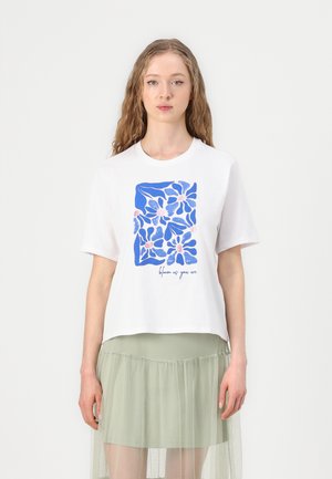 Fehér, rövid ujjú póló kék virágmintával és "bloom as you are" felirattal. Egy réteges, világoszöld tüllszoknyával kombinálva.