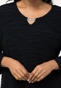 Svart topp med texturerat vågigt mönster, V-ringning med ett rhinestones accent och trekvartslånga ärmar. Slät tyg med en åtsittande design.