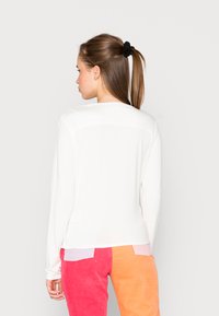 Camisola branca de mangas compridas com um decote redondo e uma ligeira bainha assimétrica. Combinada com calças de cortiça em bloco de cores em rosa e laranja.