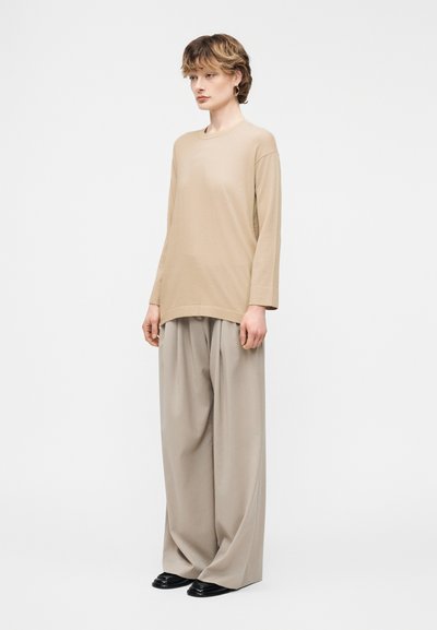 Άτομο που στέκεται φορώντας μπεζ μακρυμάνικη μπλούζα, φαρδιά ανοιχτόγρια παντελόνια και μαύρα παπούτσια μπροστά σε απλό λευκό φόντο.