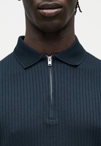 Gros plan sur une personne portant une chemise zippée côtelée bleu marine foncé avec un col texturé, montrant le bas du visage et le haut du torse.