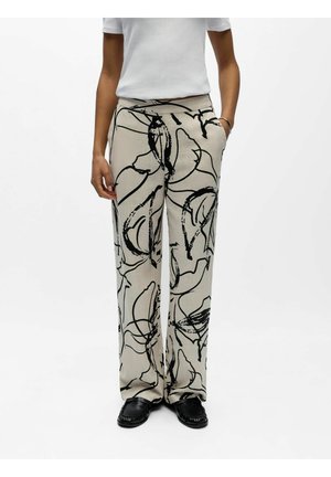 Personne portant un pantalon beige à jambes larges avec un motif abstrait en lignes noires, associé à un haut blanc à manches courtes et des mocassins noirs.