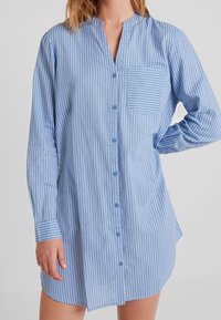 Camicia a maniche lunghe di colore azzurro chiaro con righe verticali bianche, dotata di chiusura con bottoni sul davanti, una piccola tasca sul petto e orlo arrotondato.