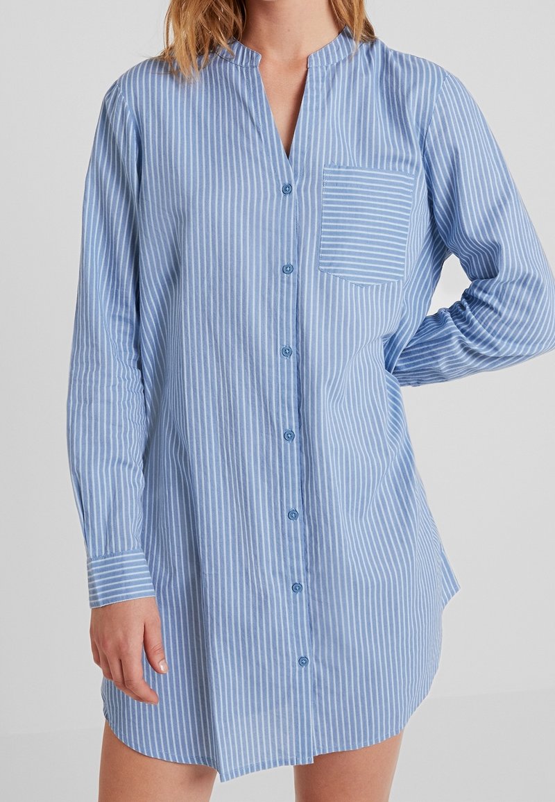 Camicia a maniche lunghe di colore azzurro chiaro con righe verticali bianche, dotata di chiusura con bottoni sul davanti, una piccola tasca sul petto e orlo arrotondato.