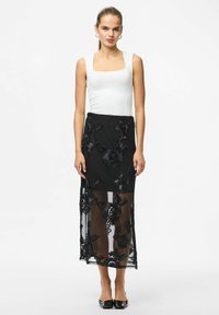 Jupe noire transparente avec broderies florales, associée à un haut blanc ajusté. Comprend une taille élastique et des éléments transparents. Chaussures plates noires portées.