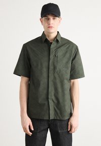 Groen overhemd met korte mouwen, voorzien van een geometrisch patroon, een borstzak, knoopsluiting en een logo-accent aan de voorkant. Gepaard met donkere jeans.