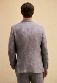 Giacca da abito in lino grigio con sottili righe verticali. Caratteristiche: taglio sartoriale, rever a lancia e bottoni sui polsini. Vista posteriore solo.
