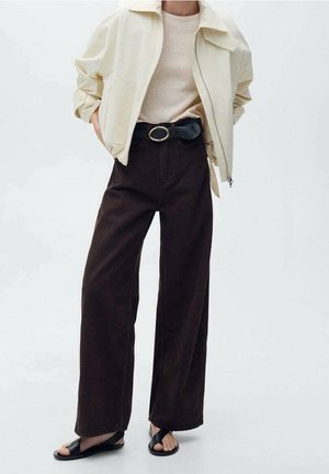 Beige leichte Jacke, cremefarbener Pullover, dunkelbraune weit geschnittene Hose, schwarzer Gürtel mit runder Schnalle und schwarze flache Sandalen. Glatte Texturen.