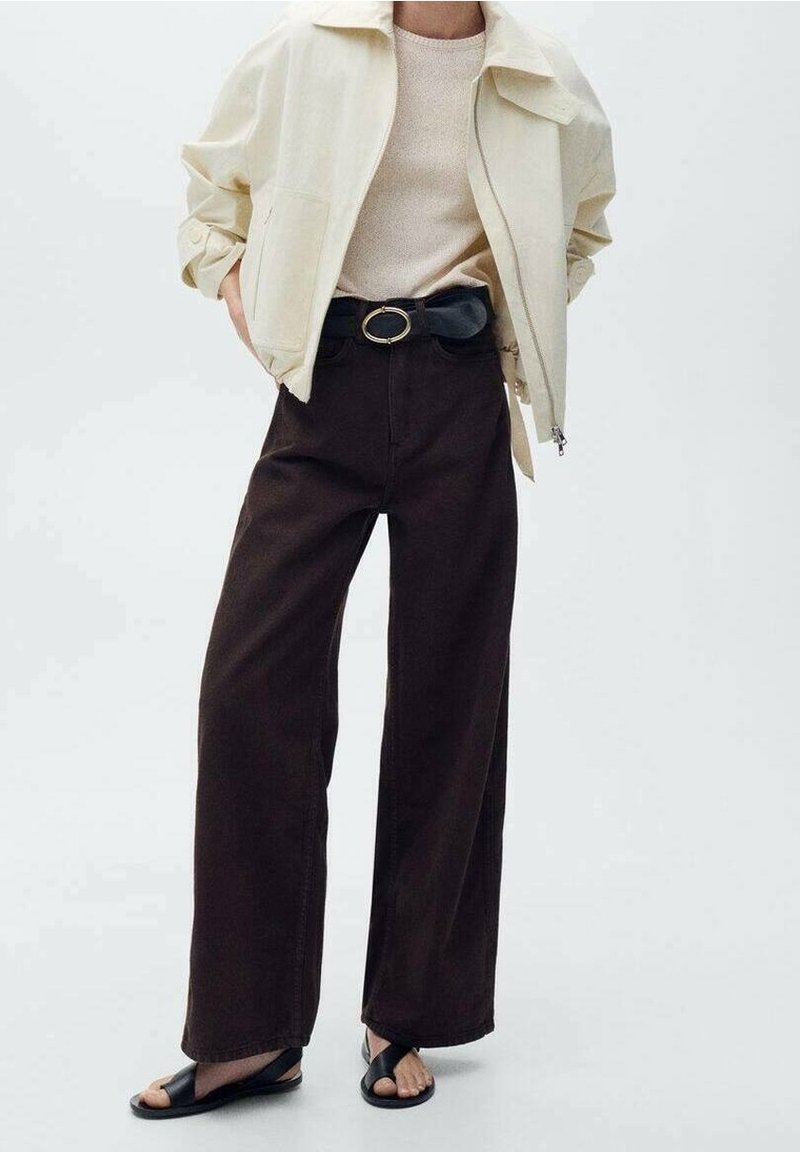 Veste légère beige, pull crème, pantalons amples marron foncé, ceinture noire avec boucle circulaire et sandales plates noires. Textures lisses.