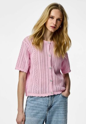 Blonde Frau, die eine hellrosa, knöpfbare Strickjacke mit kurzen Ärmeln und hellblaue, hoch taillierte Jeans trägt, steht mit einer Hand in der Tasche.