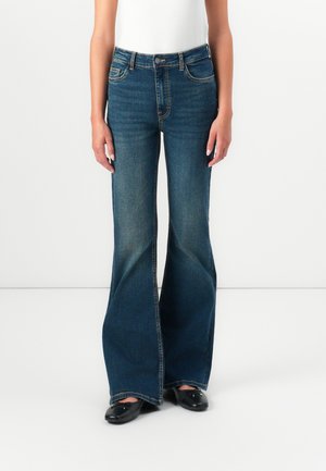 Høytlivede bukser med bredde i bena i mørk blå denim, med fem lommer, kontrastsøm og en ren finish. Flate svarte sko er på.