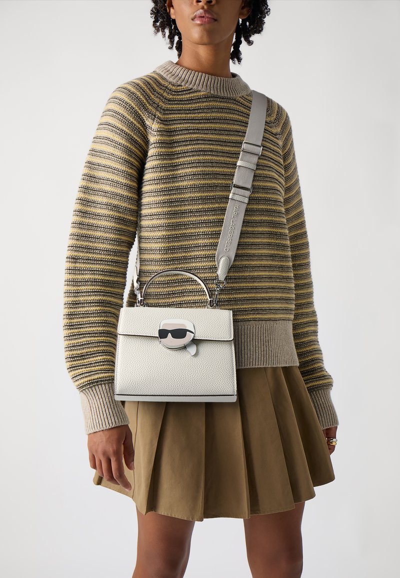 Borsa a tracolla grigia con texture, dalla forma strutturata, caratterizzata da un emblema decorativo e una tracolla staccabile, abbinata a un maglione a righe e una gonna.