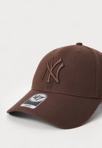 Brun hatt med brodert NY-logo, buet skjerm, tekstilstruktur og en sirkulær etikett med størrelse og merkeinformasjon.