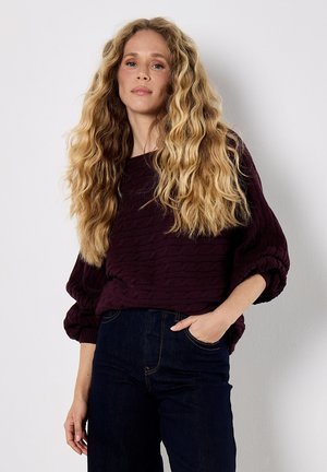 Bordeauxfarbener strukturierter Pullover mit lässigem Schnitt und Ballonärmeln, kombiniert mit dunkelblauen geraden Jeans. Der Pullover hat ein dezentes Wellenmuster.