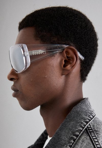 Transparent solglasögon med överdimensionerade linser, en slank design och distinkta klara bågar, uppvisade mot en texturerad grå bakgrund.