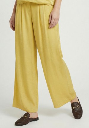 Persoon die losse gele broek en donkerbruine, getextureerde slip-on loafers met gouden accenten draagt op een effen achtergrond.