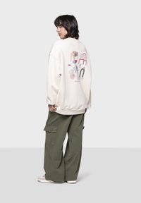 Φόρμα σε κρέμα με γραφιστικό σχέδιο στην πλάτη· συνδυασμένη με φαρδιά ελαιόχρωμα cargo παντελόνια και λευκά αθλητικά παπούτσια. Χαλαρή εφαρμογή, μαλακή υφή.