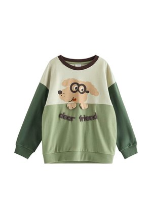 Junges Kind mit einer orangefarbenen Strickmütze, grün-beigem Sweatshirt mit einer Hundegarafik und dem Text "dear friend", blauen Jeans und weißen Sneakers.