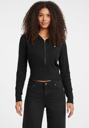 Giovane donna con capelli ricci che indossa un golfino nero a zip con lavorazione a trecce e jeans neri a vita alta su uno sfondo bianco.