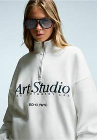 Felpa bianca con zip caratterizzata da un audace testo blu navy "Art Studio" e "SOHO//NYC". Materiale spesso con vestibilità rilassata e colletto alto. Occhiali da sole neri.