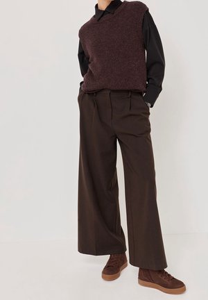 Person trägt dunkelbraune, weite Hosen, braunen, strukturierten ärmellosen Pullover über einem schwarzen Langarmshirt und braune Sneakers.