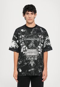 Čierne oversize tričko s podrobnou bielou kvetinovou potlačou, dizajnom vtáčej klietky a textom "Versace Jeans Couture" uprostred.