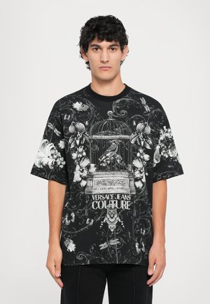 Black oversized t-shirt featuring a detailed white floral print, birdcage design, and "Versace Jeans Couture" text at the center.