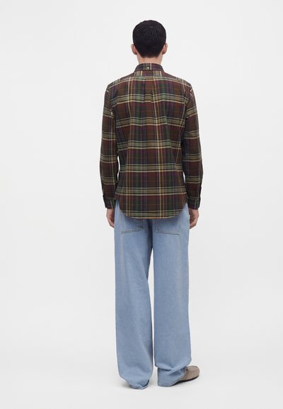 Homme debout, dos à l’objectif, portant une chemise à carreaux foncée à manches longues et un jean large bleu clair avec des chaussures marron sur fond blanc.