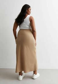 Falda maxi beige con una superficie texturizada, combinada con una blusa sin mangas blanca y zapatillas blancas, vista desde la parte posterior.
