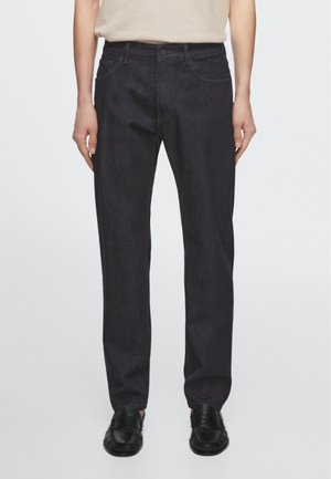 Person trägt dunkelgraue Slim-Fit-Jeans mit Fronttaschen und schwarze elegante Schuhe, stehend vor einfachem hellem Hintergrund.