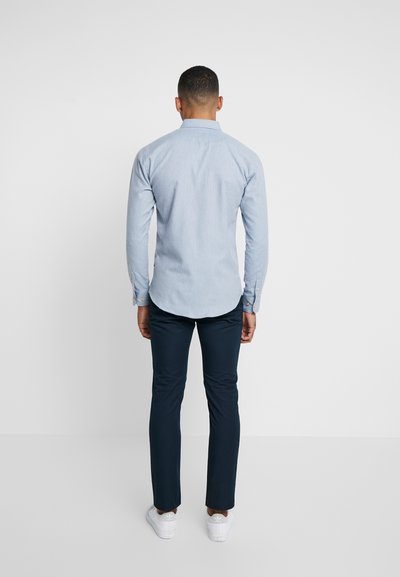 Camisa azul clara de mangas longas com colarinho abotoado, combinada com calças escuras ajustadas e ténis brancos. A vista traseira mostra a textura suave do tecido.