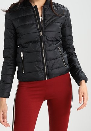 Kvinde iført en sort quiltet jakke med frontlynlås og lynlåslommer, parret med røde leggings med sidelommer.