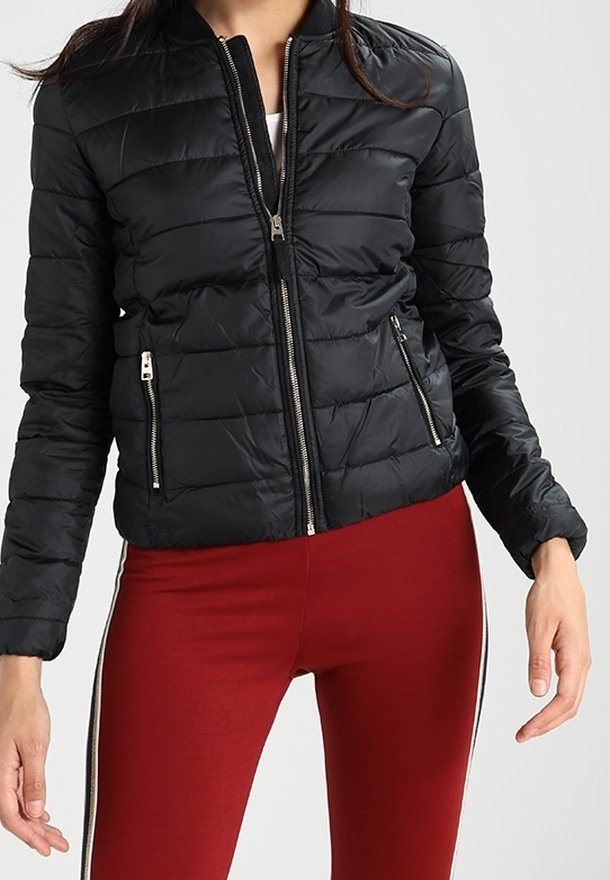 Femme portant une veste matelassée noire avec fermeture éclair frontale et poches zippées, associée à un legging rouge avec bandes latérales.