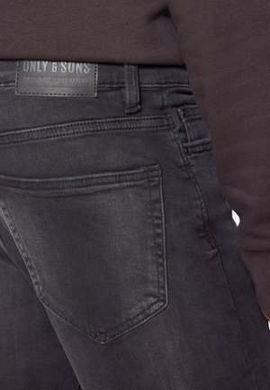 Czarne jeansy z denimu z tylną kieszenią i skórzaną naszywką z napisem "ONLY & SONS Passionate Jeans Makers", noszone z ciemną bluzką.