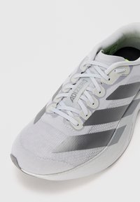 Chaussure de sport gris clair avec un dessus en tissu texturé, accents argentés et trois rayures gris foncé. Comprend des lacets plats et un col rembourré.