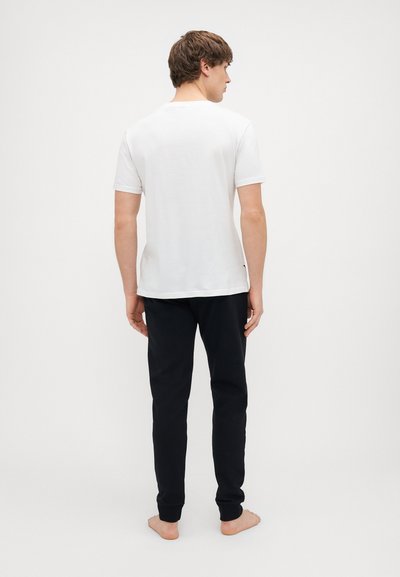 Bijela pamučna majica kratkih rukava ravnog kroja, uparena s crnim trenirkama s rebrastim manžetama, prikazana s leđa.