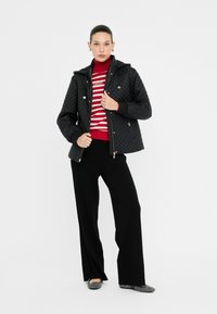 Vrouw in een zwarte gewatteerde jas over een rood-wit gestreepte coltrui, zwarte broek en geruite instappers.