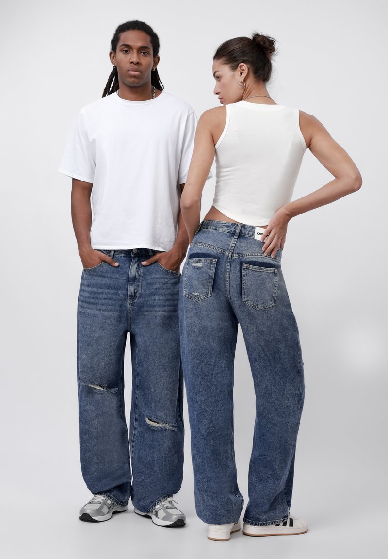 Baggy blue denim jeans with distressed details, worn by two models. One has a white tank top, and the other wears a white t-shirt.
