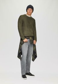 Zielony sweter dzianinowy w odcieniu oliwkowym, czapka z czarnym ściągaczem, szare dżinsy i czarne sneakersy. W pasie przewiązana jest koszula w kratkę, dodająca stylizacji szczególnego charakteru.