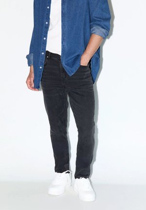 Man draagt zwarte spijkerbroek, witte sneakers, wit overhemd en een open blauwe spijkerblouse met één hand in de zak.